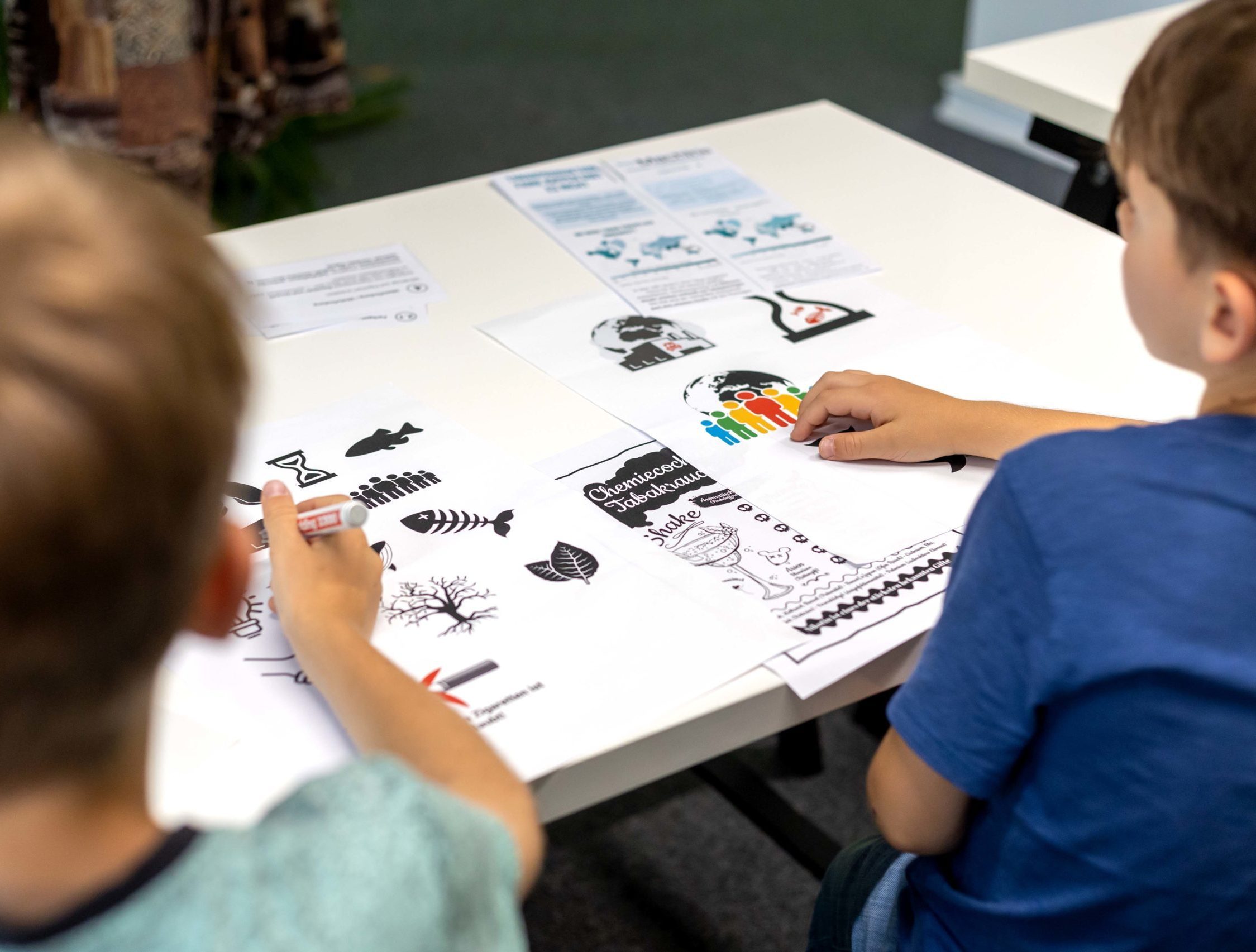 zwei Kinder sitzen am Tisch und entwerfen Logos